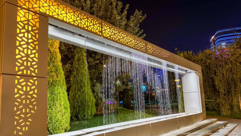 Illuminated rectangular frame with gold geometric trim and a curtain of falling water at night.