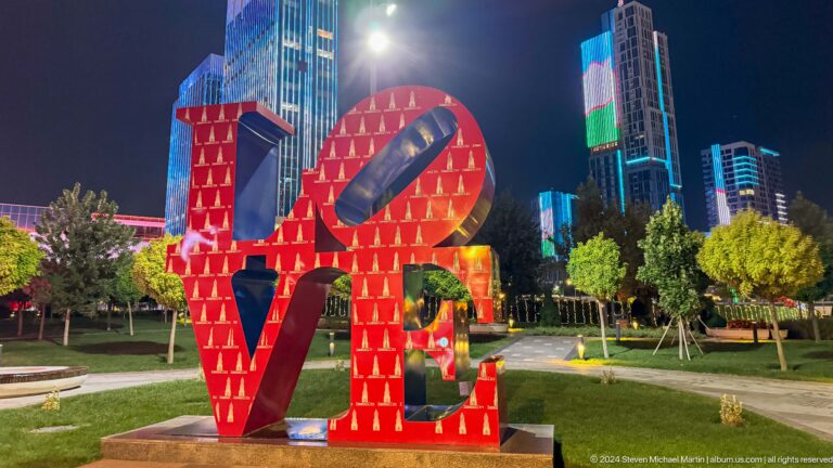 Large red LOVE sculpture on a lawn with illuminated high-rise towers behind at night.