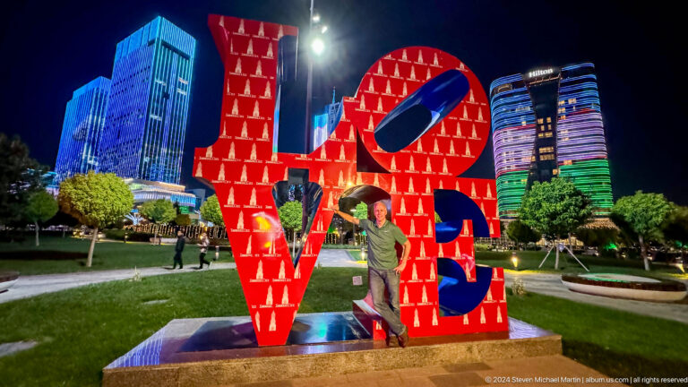 Man posing inside a red LOVE sculpture with Hilton and blue-lit towers behind at night.