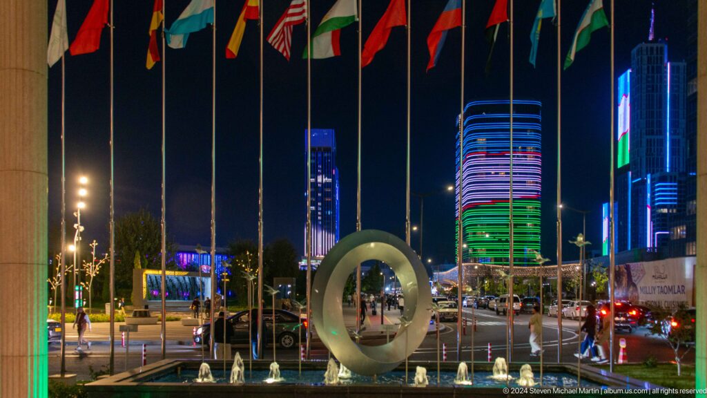 Night plaza with international flags, a circular metal sculpture, small fountains, pedestrians, and brightly illuminated towers.
