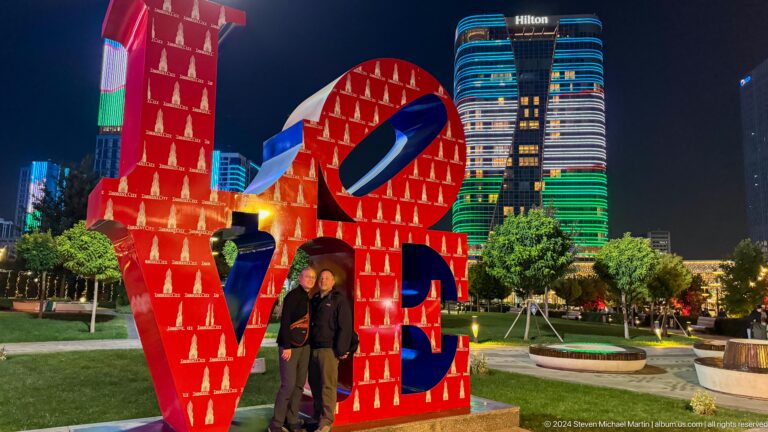 Two people posing inside a red LOVE sculpture with the illuminated Hilton tower behind.
