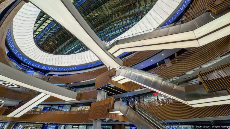 Upward view inside a mall atrium with a glass roof, oval light ring, escalator, and wood-clad balconies.