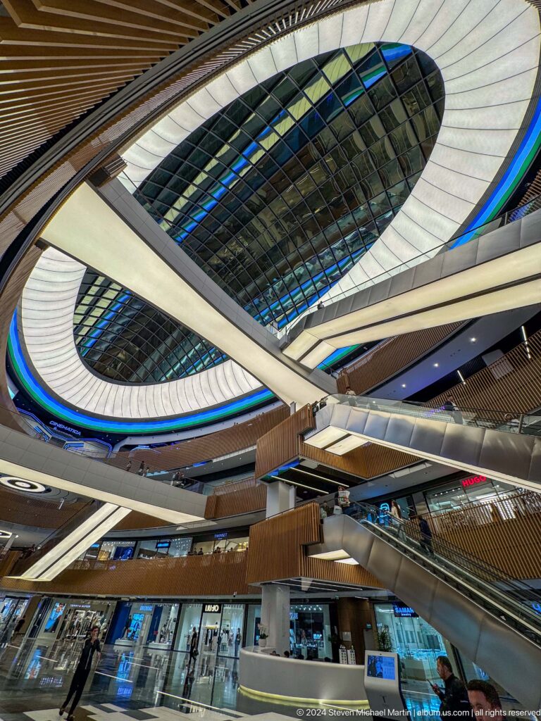 Upward mall view of an oval glass roof, blue accent bands, a diagonal escalator, and wood balconies.