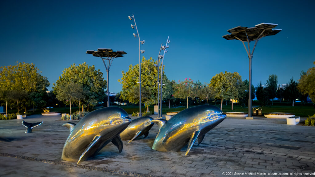 Three polished metal dolphin sculptures rising from a paved plaza with trees behind.
