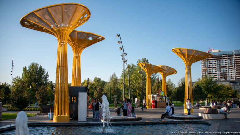 Visitors among yellow tree-like canopies, circular benches, and fountains in a sunny park plaza.