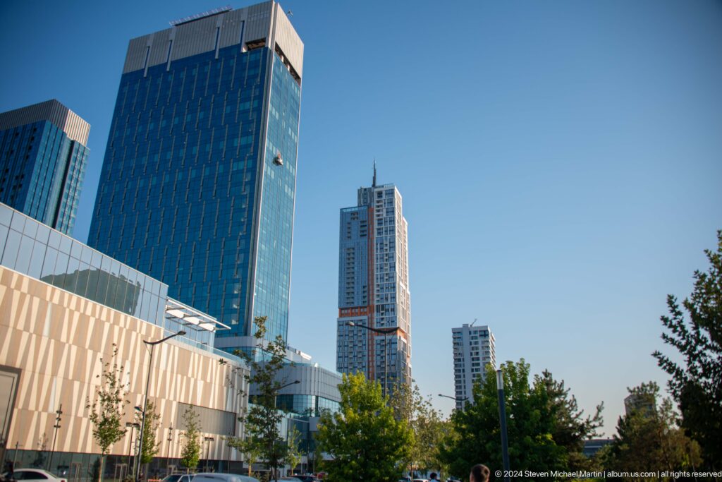 Daytime street view with a pale mall facade, trees, and several modern high-rise towers under blue sky.