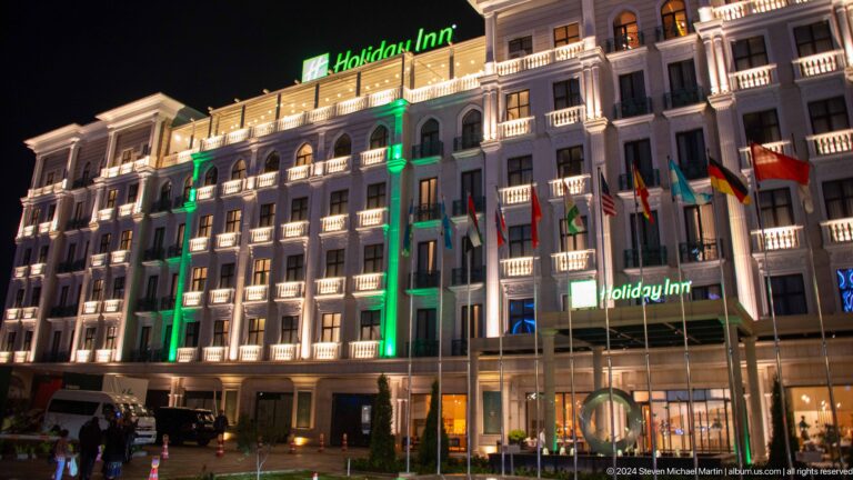 Night view of the Holiday Inn Tashkent City facade with arched windows, balconies, white outline lights, and green illumination.
