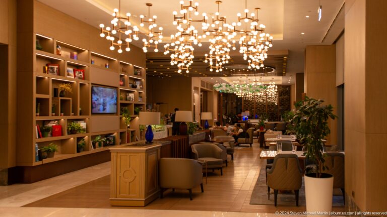 Long hotel lounge with bookshelves, seating, plants, televisions, and branching globe pendant lights.