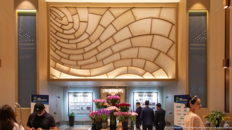 Large cream padded wave-pattern wall above Wall of Fame displays, flowers, and people in a hotel.