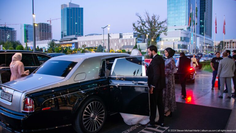 Uniformed attendant opening a black luxury sedan for a woman outside a hotel at dusk.
