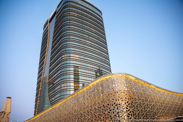 Rounded glass hotel tower behind a curving gold geometric lattice roof in daylight.