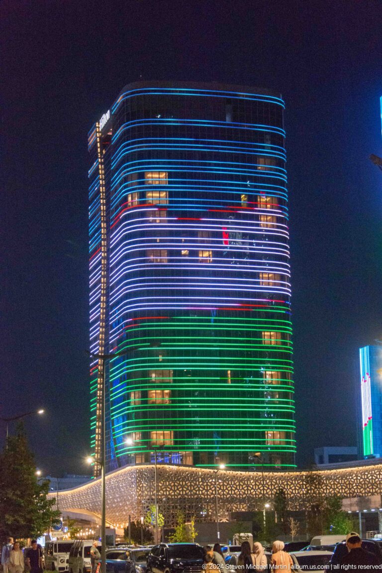 Two illuminated high-rise buildings at night, one pale white and one glowing red, above low structures.