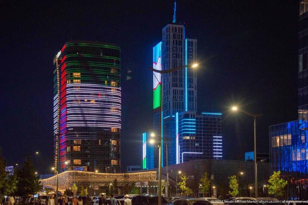 Cars travel along a wide road at night beneath blue-, green-, and red-lit high-rise buildings.
