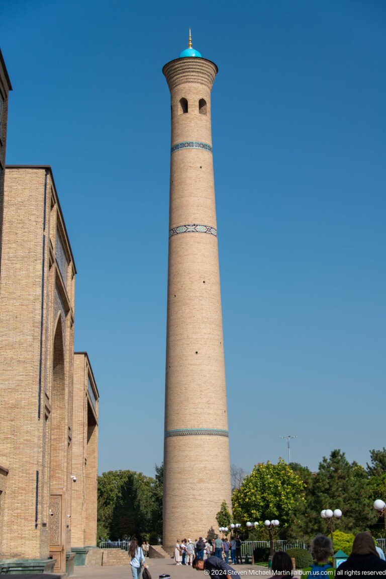 Vertical pale-brick minaret beside a masonry facade against blue sky at the Khast Imom complex in Tashkent.