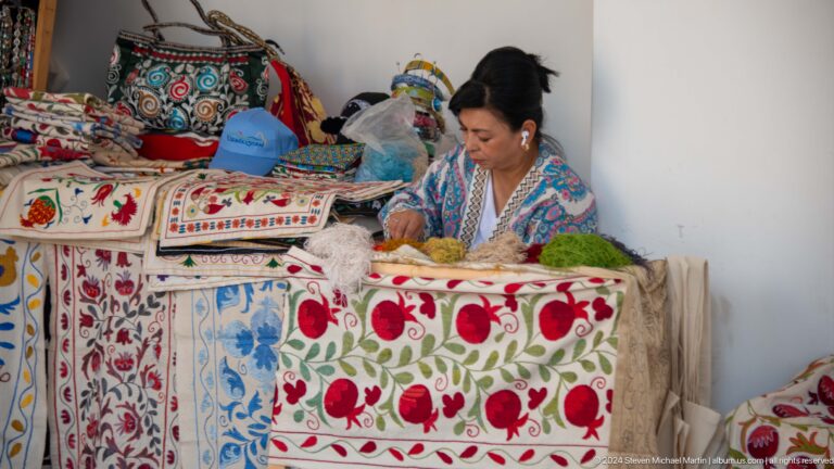Seated woman sewing among vivid suzani-style embroidered textiles at the Khast Imom complex in Tashkent.