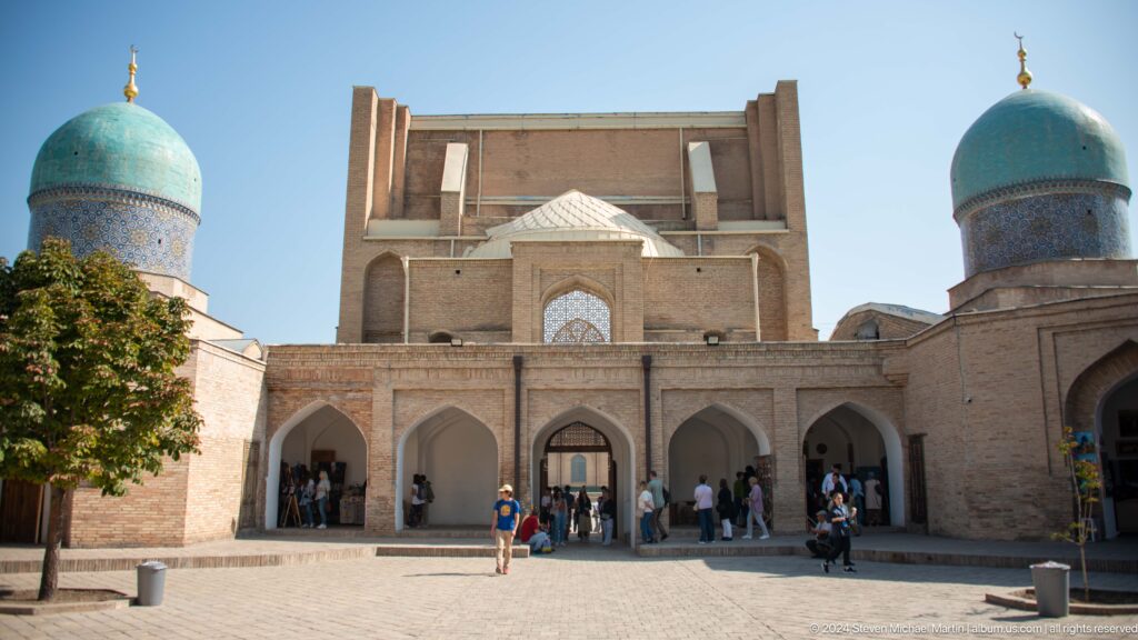 Deep brick entrance with upper recessed arch, arcaded ground floor, and visitors at the Khast Imom complex in Tashkent.