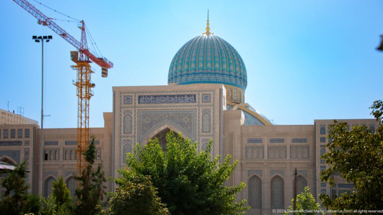 Large pale monumental facade with high central arch, blue dome, trees, and crane at the Khast Imom complex in Tashkent.