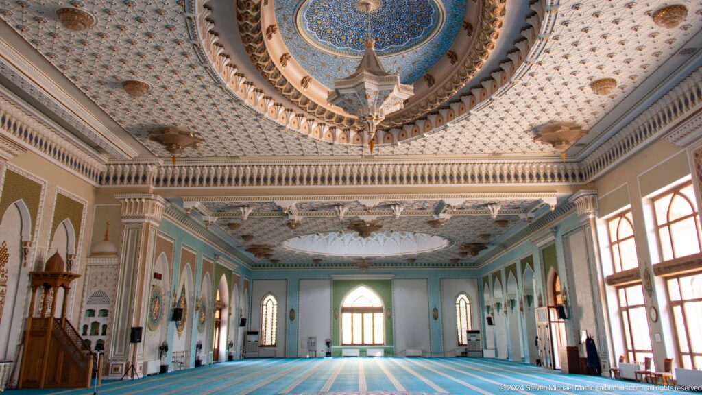 Wide prayer hall with pale blue walls, arched windows, carpets, chandelier, and decorated ceiling at the Khast Imom complex in Tashkent.