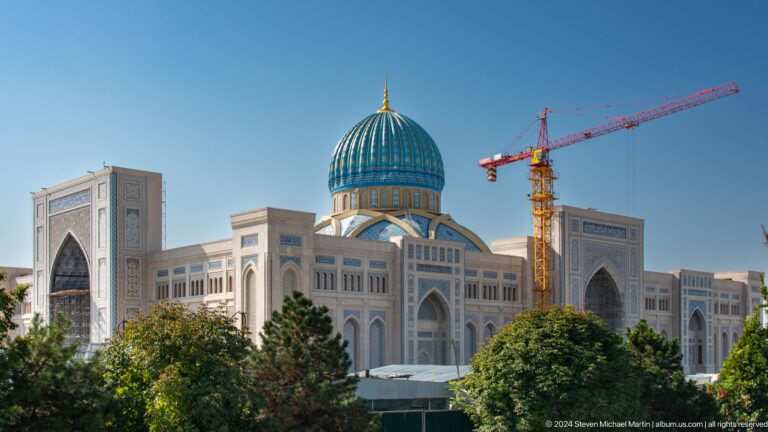 Large pale facade and blue dome partly obscured by trees and construction crane at the Khast Imom complex in Tashkent.