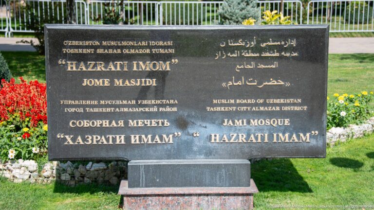 Multilingual black sign reading hazrati imom / hazrati imam in landscaped grounds at the Khast Imom complex in Tashkent.