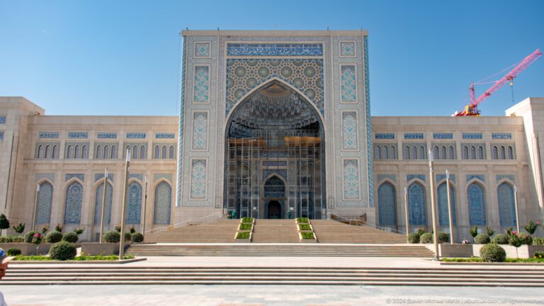 Frontal view of vast pale facade with high central tiled arch and formal steps at the Khast Imom complex in Tashkent.