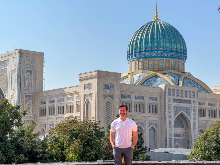 Man in white shirt and cap posed against pale building and blue dome at the Khast Imom complex in Tashkent.