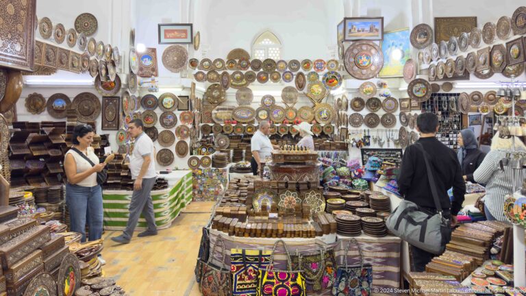 Dense stand of brass plates, ceramics, textiles, and small decorative objects with shoppers at the Khast Imom complex in Tashkent.