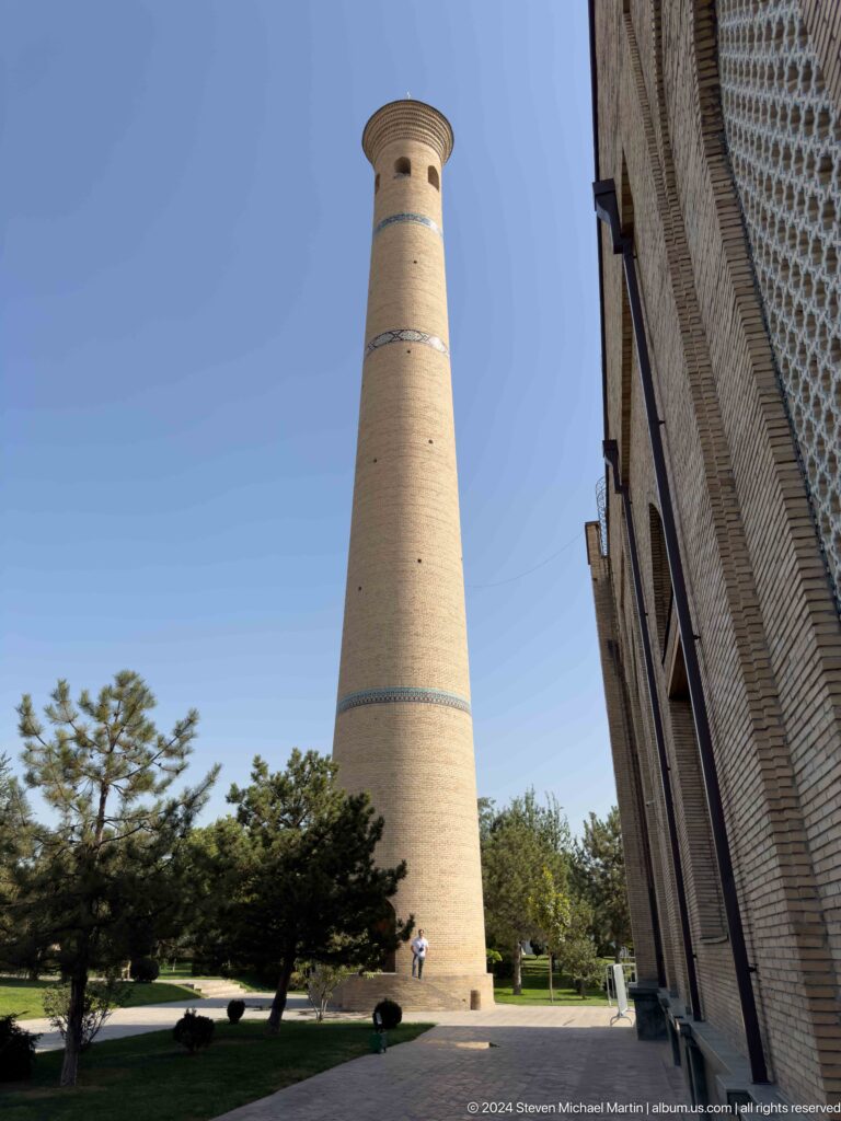 Tall pale-brick minaret rising beside a shaded building edge and trees at the Khast Imom complex in Tashkent.