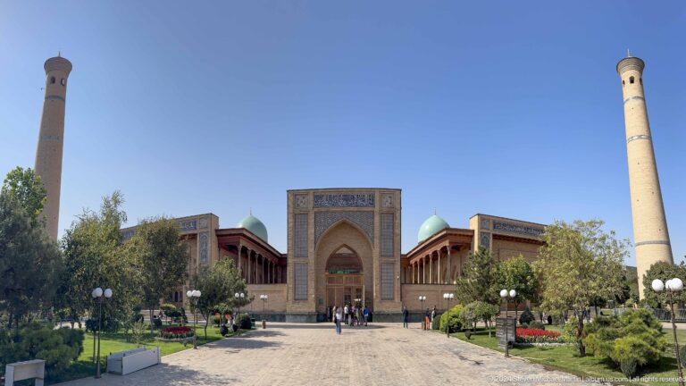 Distant tiled portal, turquoise domes, trees, and pedestrian path under clear sky at the Khast Imom complex in Tashkent.