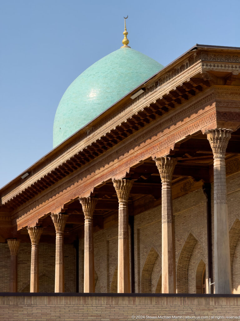 Row of carved wooden columns beneath deep decorated eaves against brick wall at the Khast Imom complex in Tashkent.