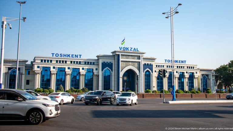 Tashkent railway station with blue windows, pointed central entrance, Uzbek flag, and traffic in front.