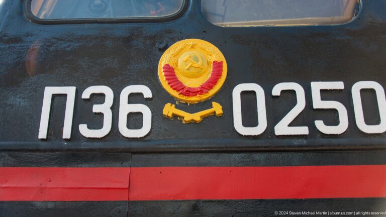 White locomotive number 736-025 beside a gold-and-red Soviet emblem on black metal.