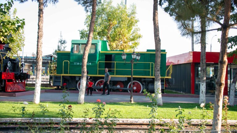 Green diesel shunting locomotive with yellow railings and red wheels behind trees at the museum.