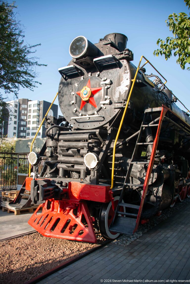 Oblique front of a black steam locomotive with Soviet star, lamps, red beam, pipes, and ladder.
