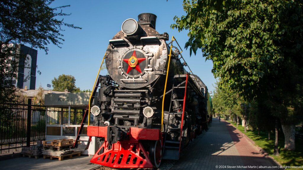 Head-on black steam locomotive with red pilot, Soviet star, lamps, and a tree-lined path.