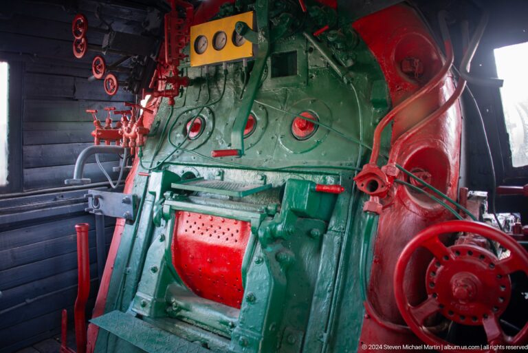 Green steam locomotive backhead with gauges, red valves, pipes, handwheel, and firebox door.
