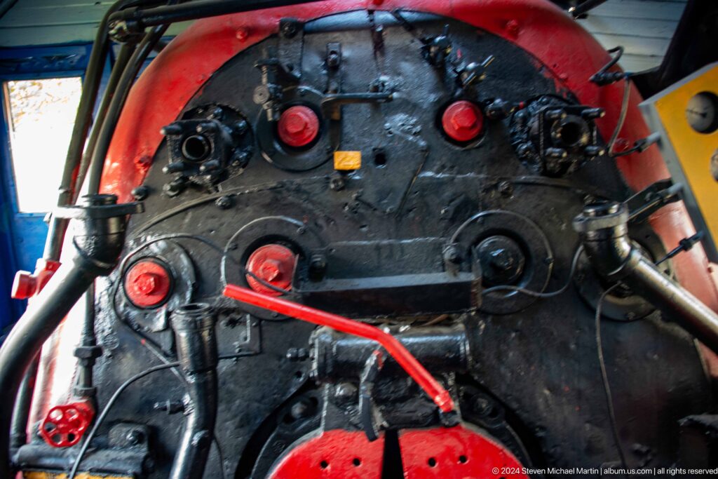 Black locomotive boiler backhead covered with red valves, pipes, round covers, and bolted fittings.