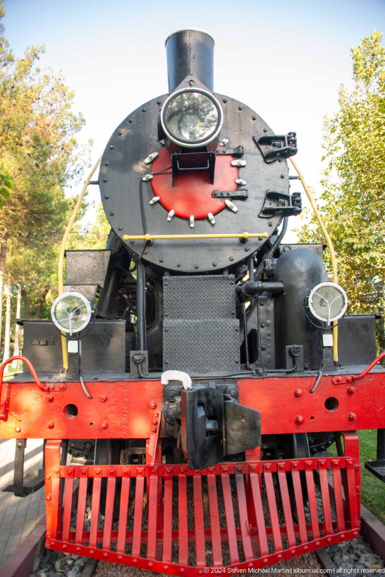 Close front of a steam locomotive with two lamps, mesh grille, coupling, and red buffer beam.