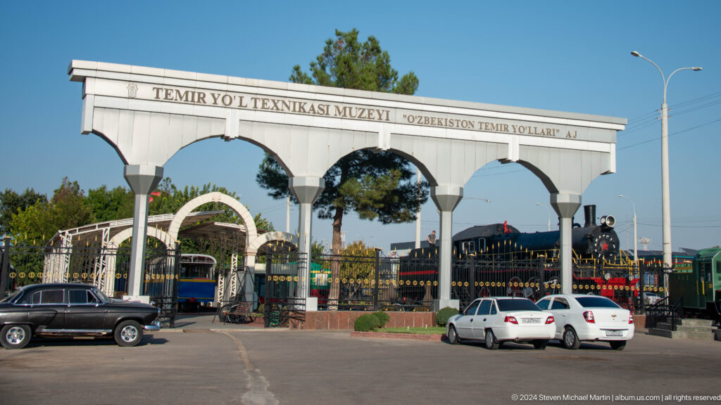White arched entrance to the Tashkent Railway Museum with locomotives visible behind decorative gates.