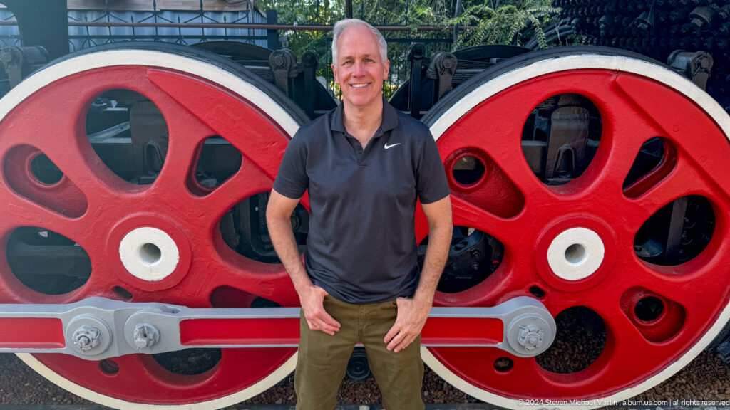 Male visitor standing between two giant red locomotive wheels with a silver connecting rod behind him.