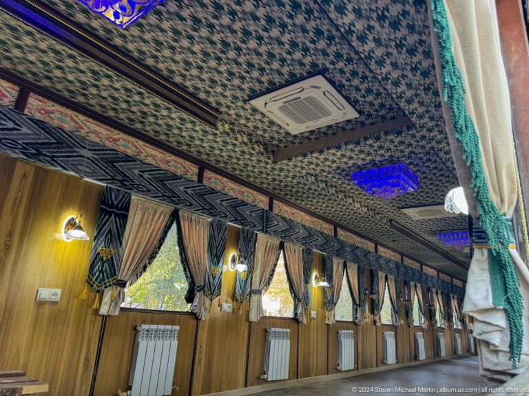 Wood-paneled railway carriage interior with patterned ceiling, curtains, wall lamps, and blue accent lighting.
