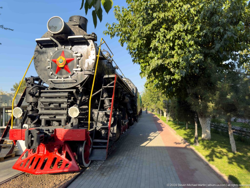 Black steam locomotive with red pilot and Soviet star beside a leafy paved museum walkway.