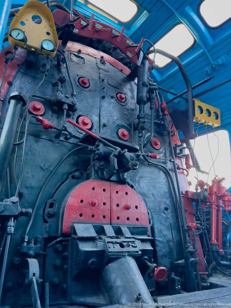 Blue-lit steam locomotive cab with dark boiler backhead, red valves, pipes, and firebox opening.
