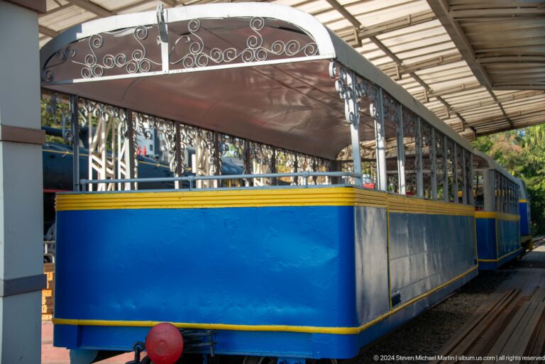 Blue and yellow open-sided passenger railcar with decorative metalwork beneath a shelter.