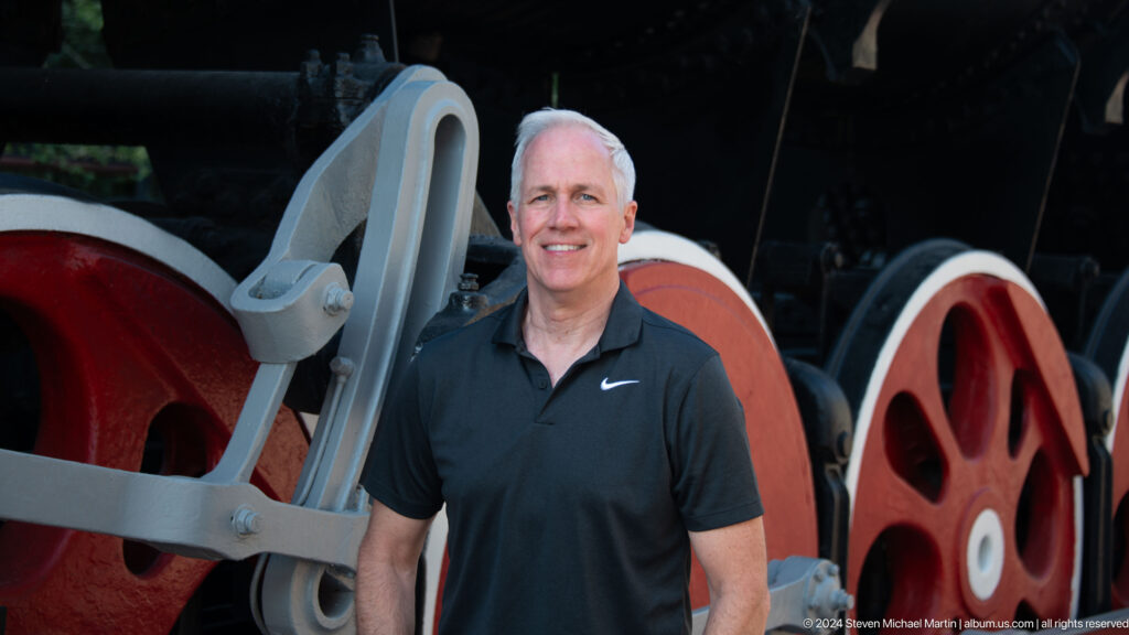 Smiling male visitor standing beside a gray connecting rod and large red locomotive wheels.