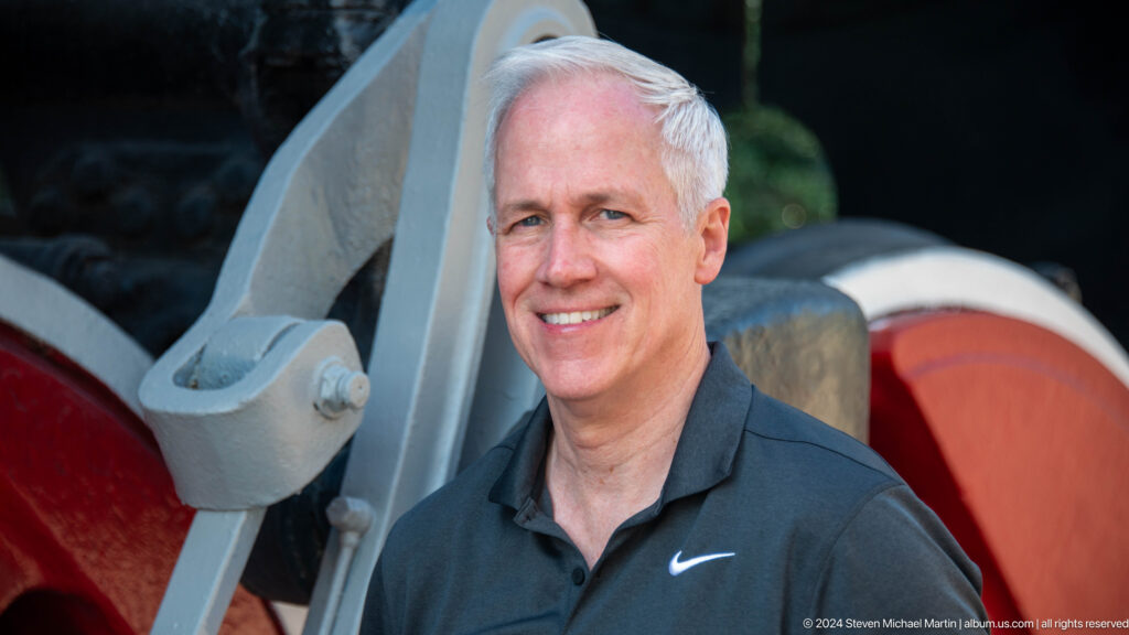 Close portrait of a smiling male visitor with locomotive linkage and a red wheel behind him.