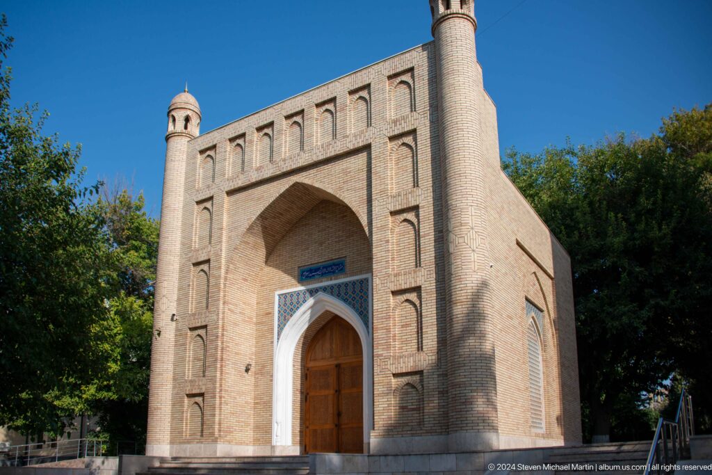 Brick mausoleum facade with deep pointed portal, carved wooden door, blue inscription panel, and corner turrets.