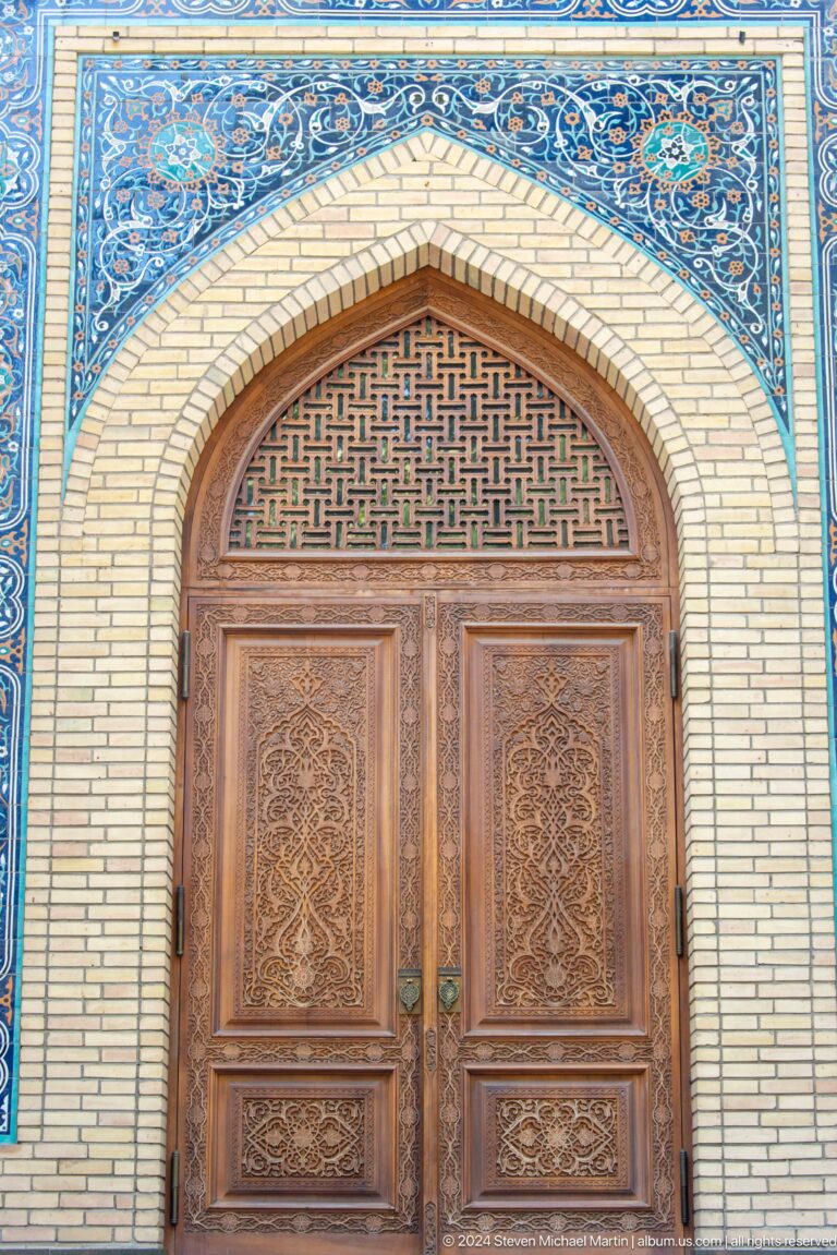 Carved wooden double door beneath a geometric lattice transom in a pointed blue-edged arch.