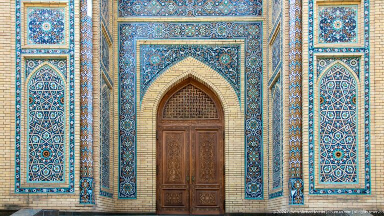 Pointed wooden door framed by blue geometric and floral tile panels on a tan brick facade.