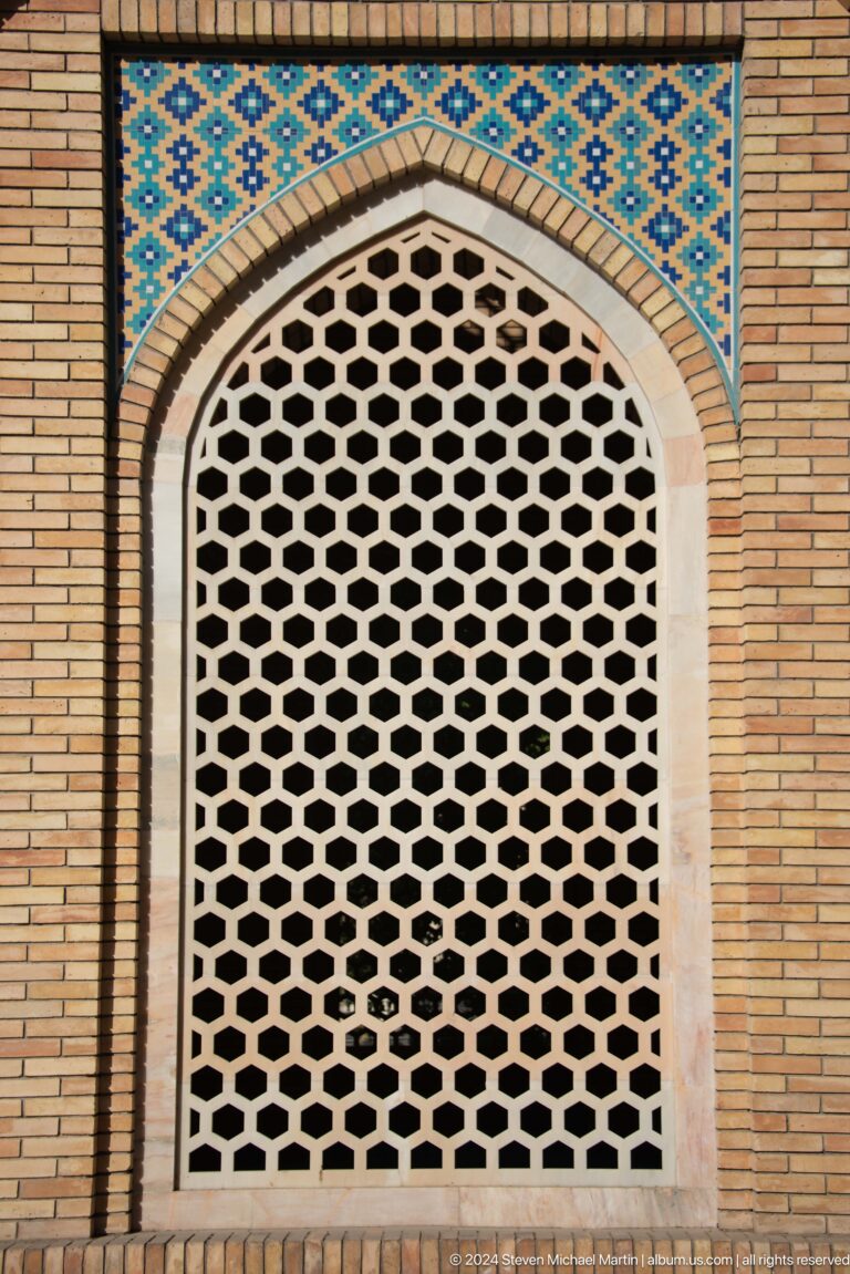 White hexagonal lattice screen filling a pointed opening in a tan brick wall.
